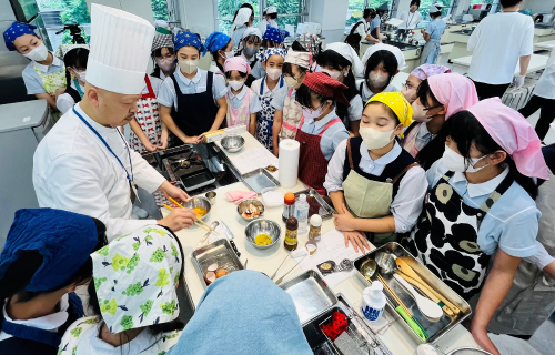 調理室で、エプロンと三角ナプキンをつけた生徒たちが、食材の載った調理台を囲みながら、コック姿の講師の説明を聞いている